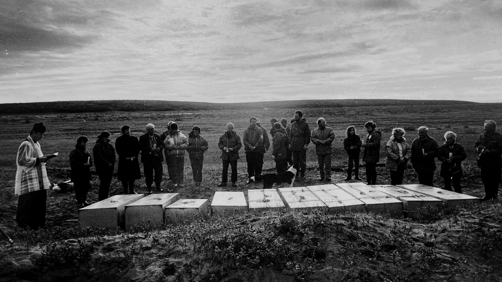 A line of caskets with people gathered behind them, in archival footage shown in MONUMENT.