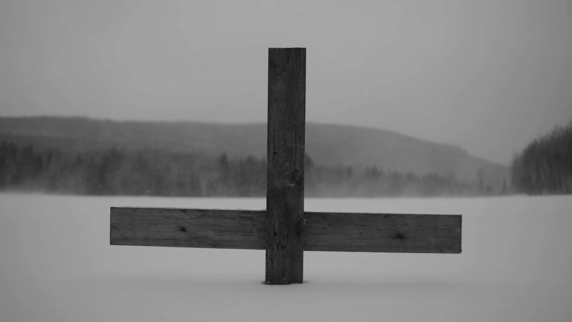 A wooden cross in the snow with mountains beyond, in MONUMENT.