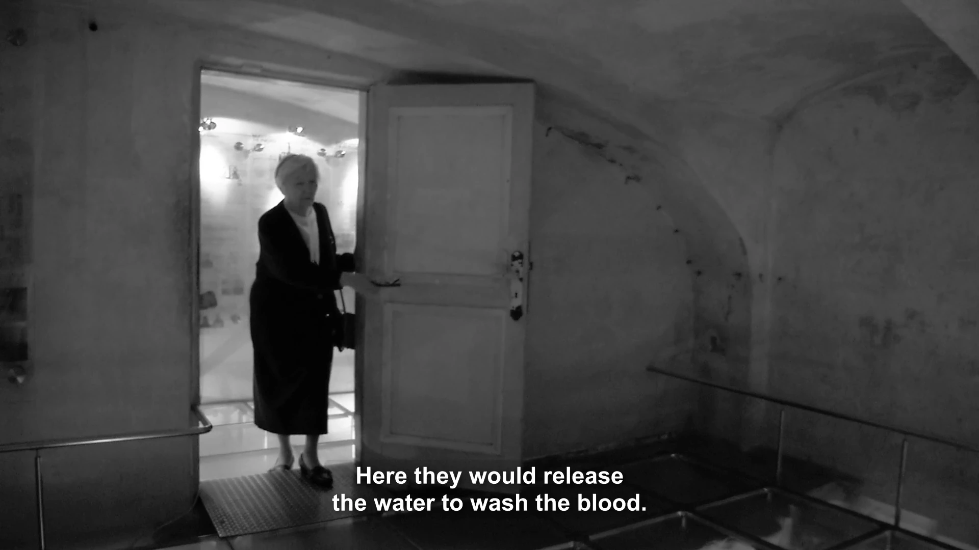 A woman standing in a prison doorway in MONUMENT; the film subtitle describes washing the floor.