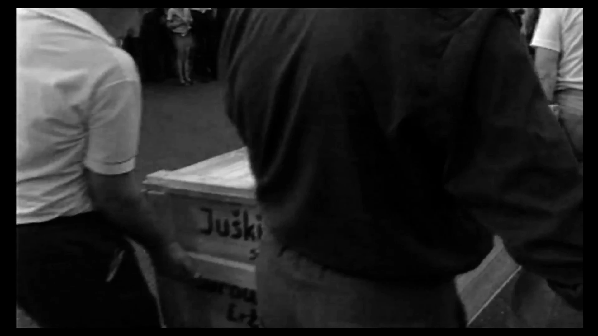 People carrying a casket at an airport, in footage shown in MONUMENT.