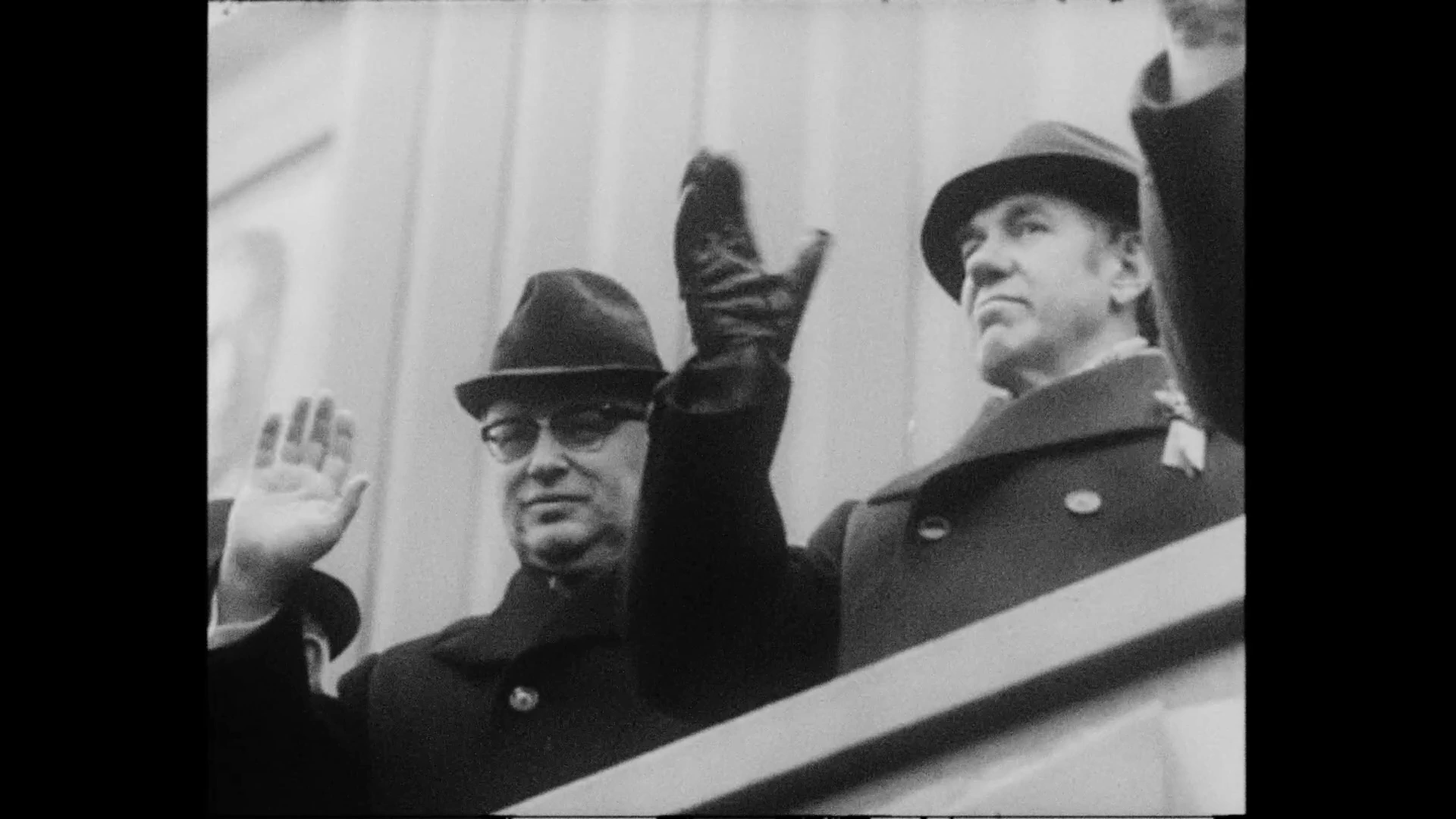 Officials waving from a balcony in archival footage shown in MONUMENT.